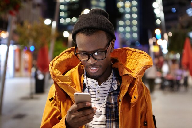 Tiro na cabeça do jovem estudante afro-americano bonito andando pela cidade à noite, segurando o telefone móvel, usando wifi urbano, navegando em fotos nas mídias sociais. Comunicação e tecnologia moderna