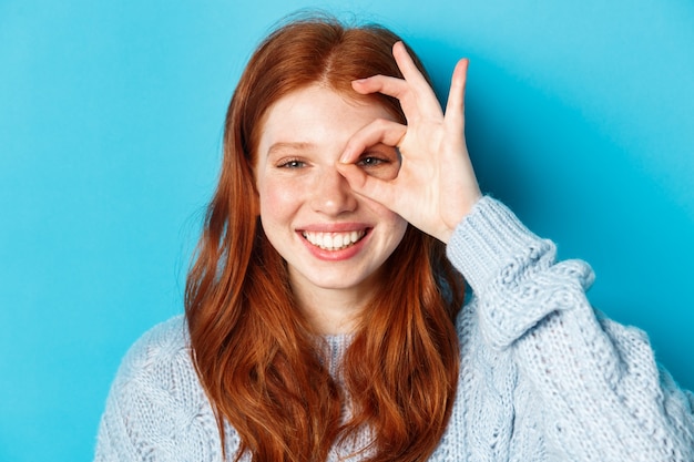 Tiro na cabeça do modelo feminino ruiva alegre mostrando sinal bem sobre o olho, sorrindo satisfeito e feliz, em pé contra um fundo azul.