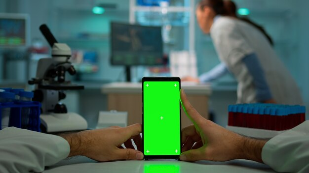 Tiro POV de químico usando smartphone com tela verde em laboratório biológico. Trabalhador médico vestindo jaleco branco na clínica trabalhando com celular com chroma key em exibição isolada em laboratório médico