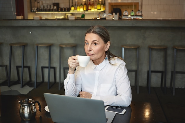 Tiro sincero de empresária madura pensativa em camisa formal, apreciando o café durante o almoço, sentado no café com laptop genérico e celular de tela em branco na mesa. negócios, idade e tecnologia