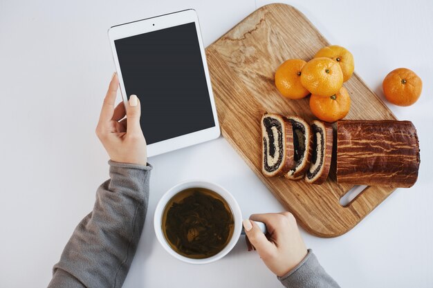 Tiro superior das mãos femininas segurando o tablet digital enquanto tomando café da manhã. Mulher inteligente aumenta a energia com uma xícara de chá e comer tangerinas com bolo enrolado, me sentindo bem na manhã de inverno.