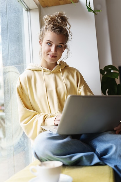 Tiro vertical da bela mulher sorridente usando laptop enquanto está sentado na janela com o laptop no colo.