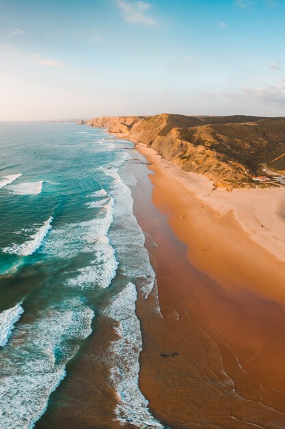 Tiro vertical das ondas do mar de tirar o fôlego e da praia com falésia rochosa sob o céu azul