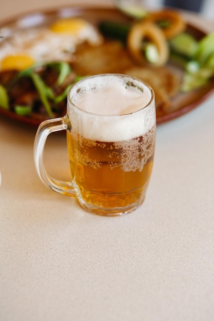 Tiro vertical de copo de cerveja gelada espumoso em pé perto de prato de aperitivos cerveja fresca leve com espuma alta espessa em pé na superfície de madeira lisa da mesa do restaurante ou pub olhando na boca