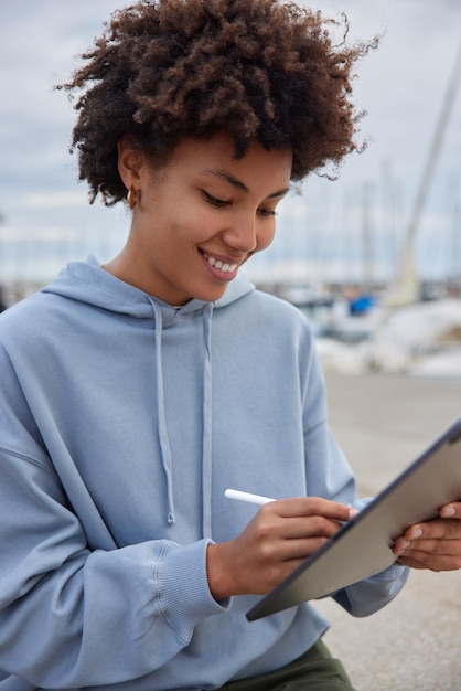 Foto grátis tiro vertical de feliz designer feminino criativo trabalha com tablet e caneta ao ar livre, vestido de moletom azul tem um sorriso no rosto feliz por terminar o trabalho. estudante universitário prepara projeto online