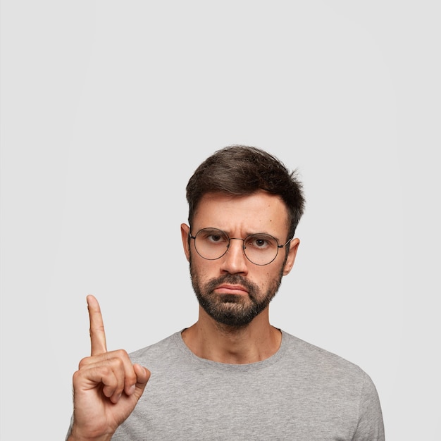 Tiro vertical de jovem com expressão de desagrado grave, restolho grosso, cabelo escuro, pontas com o dedo indicador para cima, vestido casualmente, isolado sobre parede branca. veja isso!