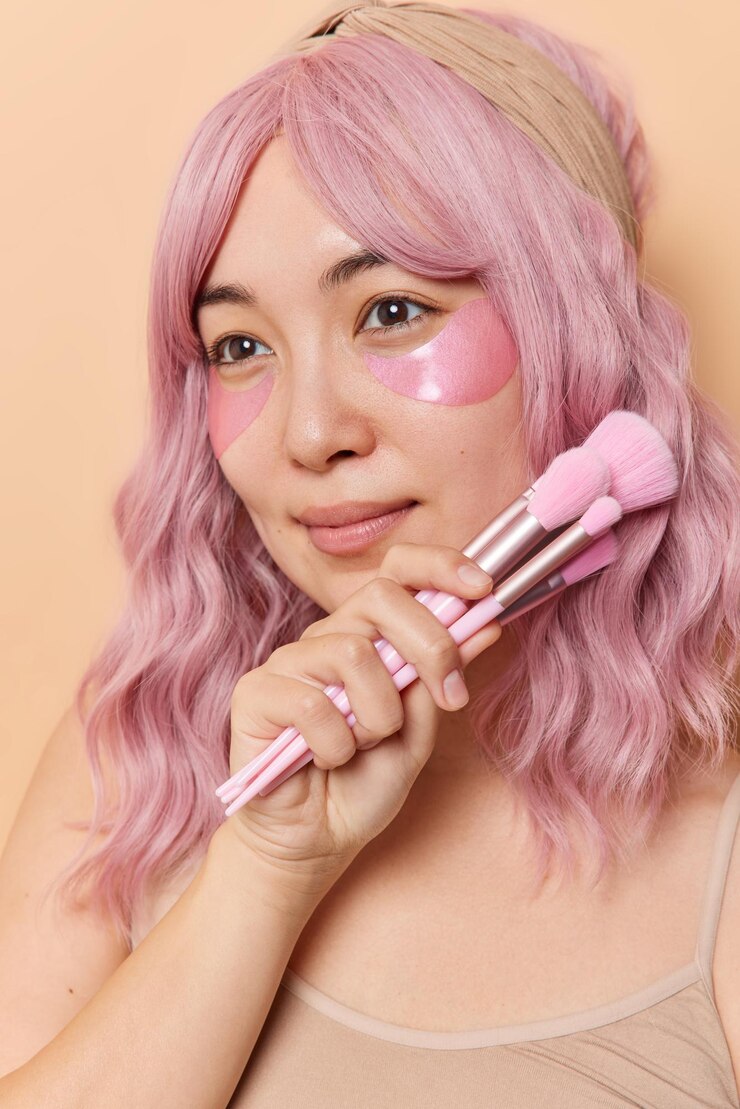 Foto grátis tiro vertical de mulher asiática de cabelos rosa pensativa segura escovas cosméticas perto do rosto desvia o olhar aplica almofadas de hidrogel sob os olhos para rejuvenescimento isolado sobre fundo bege rotinas de cuidados com a pele