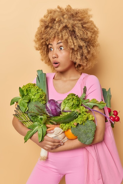 Foto grátis tiro vertical de mulher chocada impressionada carrega saco de tecido de legumes frescos retorna do mercado descobre notícias impressionantes vestidas com roupas rosa isoladas sobre fundo bege entrega de alimentos