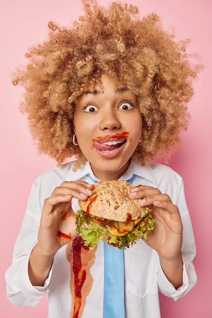 Foto grátis tiro vertical de mulher de cabelos cacheados gosta de comer junk food lambe os lábios manchados com ketchup segura hambúrguer grande tem menu de fast food aluna tem dia de refeição de trapaça sente fome depois das palestras