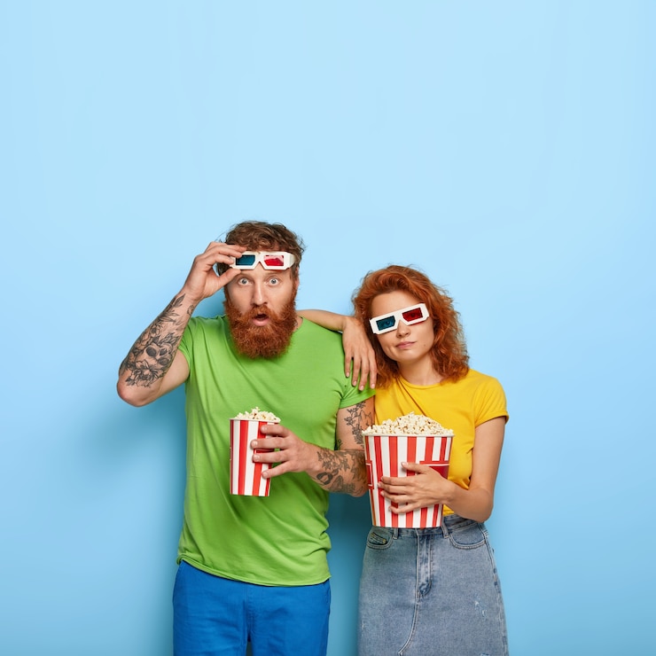 Foto grátis tiro vertical de um homem chocado encara com surpresa, tira os óculos 3d, mulher entediada e triste se inclina para o ombro, passa o tempo livre no cinema, come pipoca saborosa, pose interior. pessoas e entretenimento