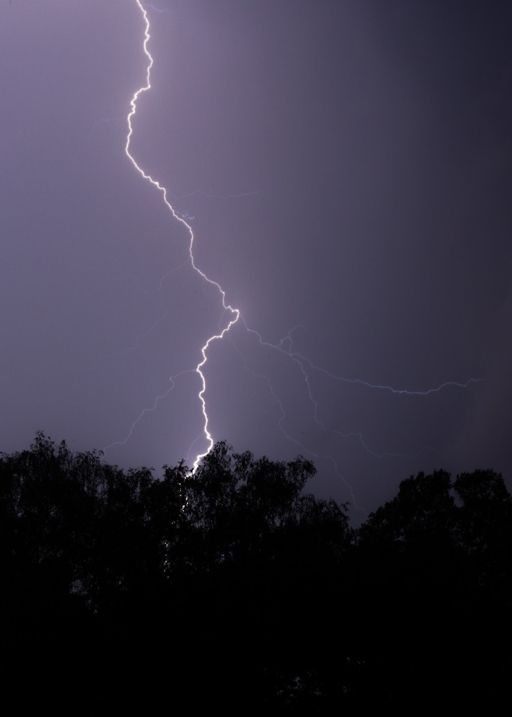 Foto grátis tiro vertical de um raio atingindo uma árvore à noite com um céu roxo e árvores na frente