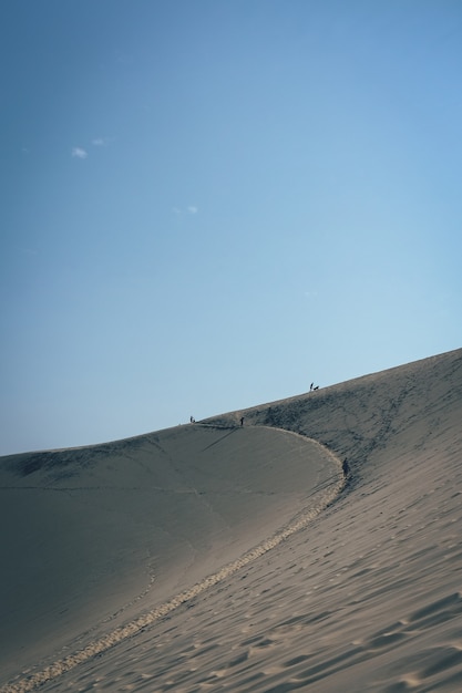 Tiro vertical de uma duna de areia com pessoas andando à distância e um céu azul claro