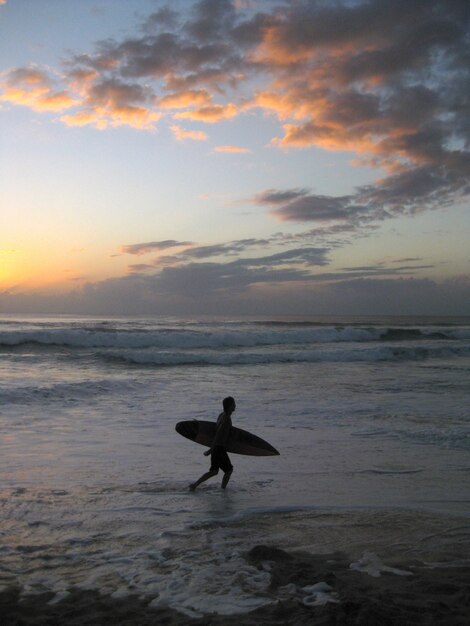 Tiro vertical de uma pessoa segurando uma prancha andando perto de um mar ondulado durante o pôr do sol