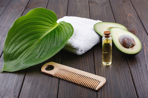 Toalha branca, abacate, garrafa de vidro pequena e pente de madeira para cabelo deitar em uma mesa de madeira