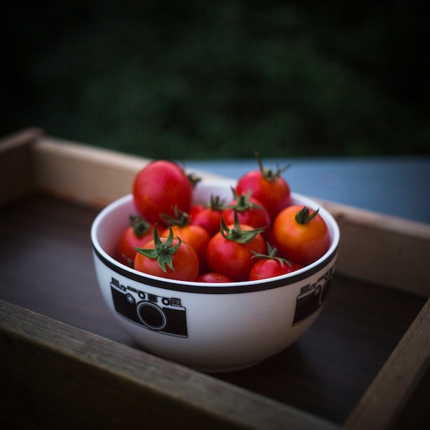 Tomates-cereja maduros vermelhos em tigela branca na bandeja de madeira