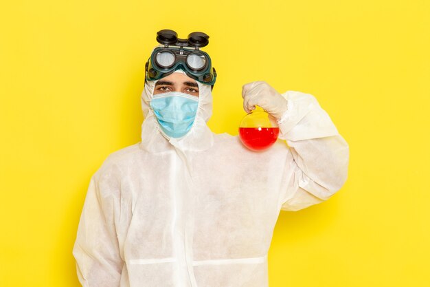 Trabalhador científico masculino de vista frontal em traje de proteção especial segurando o frasco com solução vermelha na superfície amarela