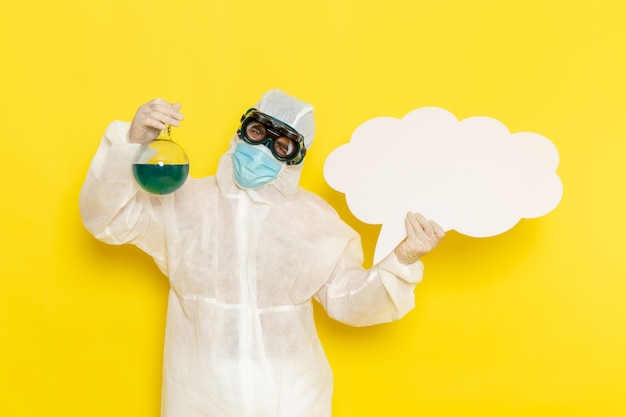 Trabalhador científico masculino de vista frontal em um terno especial segurando um frasco com uma solução verde e uma grande placa branca na mesa amarela