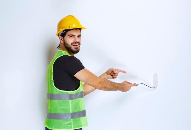 Trabalhador da construção civil com um capacete amarelo pintando a parede branca com um rolo aparador.