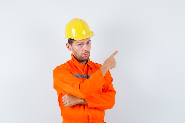 Trabalhador da construção civil de uniforme, capacete apontando para o canto superior direito e olhando confiante, vista frontal.