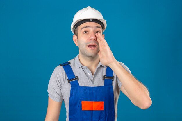 Trabalhador da construção civil em uniforme e capacete de segurança, gritando algo e mantendo as mãos perto de sua boca aberta no azul isolado