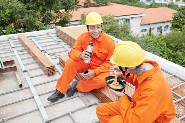 Trabalhador da construção civil em uniforme laranja, bebendo café quente e olhando para o colega almoçando quando ...