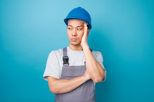 Trabalhador da construção civil jovem pensativo usando capacete de segurança e uniforme, mantendo a mão na cabeça olhando para o lado