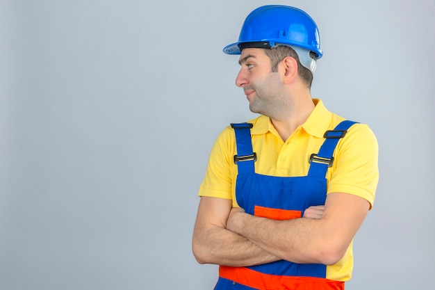 Trabalhador da construção civil no capacete de segurança uniforme e azul em pé com as mãos cruzadas, olhando para longe para o lado em branco, com espaço de cópia