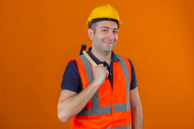 Trabalhador da construção civil usando colete e capacete de segurança amarelo, segurando o martelo com um sorriso no rosto, isolado na laranja
