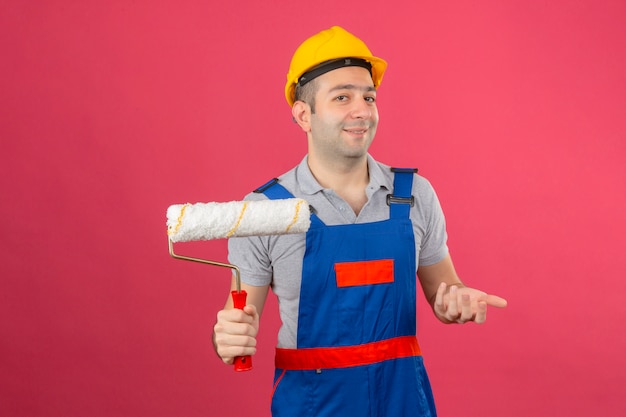 Foto grátis trabalhador da construção civil usando uniforme e capacete de segurança fazendo gesto confuso com a mão e expressão como pergunta segurando o rolo de pintura isolado na rosa