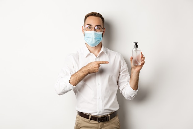 Trabalhador de escritório com máscara médica apontando para desinfetante de mão, mostrando fundo branco, anti-séptico.