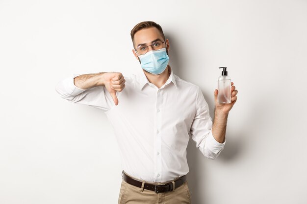 Trabalhador de escritório com máscara médica descontente, mostrando desinfetante para as mãos e o polegar para baixo, em pé