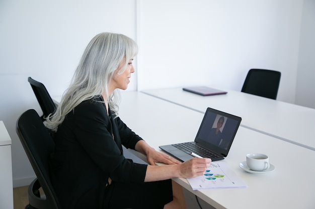 Trabalhador de escritório feminino amigável conversando com um colega via chat de vídeo no laptop enquanto está sentado à mesa com uma xícara de café e analisando o diagrama. Conceito de comunicação online