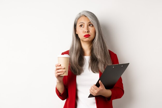 Trabalhador de escritório feminino asiático cansado segurando a prancheta e o copo de papel, bebendo café e expirando com o rosto exausto, em pé sobre um fundo branco.