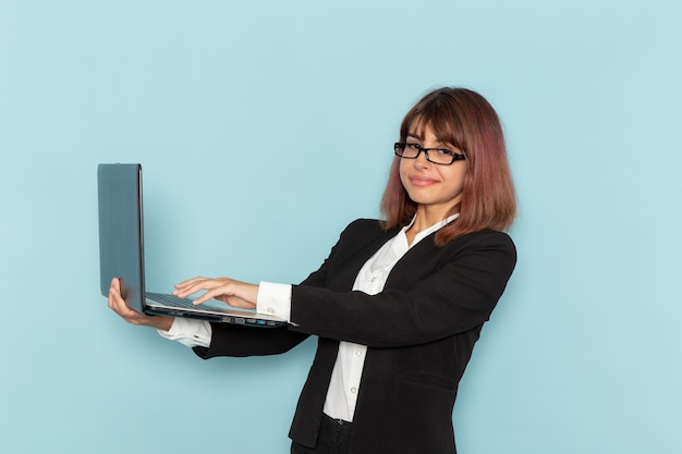 Trabalhador de escritório feminino em terno estrito sorrindo e usando o laptop na superfície azul.