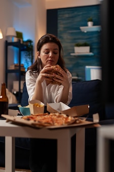 Trabalhador de escritório sentado no sofá olhando saboroso hambúrguer delicioso na sala de estar em frente à televisão. mulher de pé à mesa com pizza grande de refeição de fast-food para viagem.
