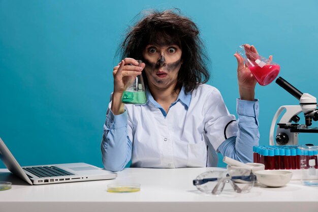 Trabalhador de laboratório louco olhando bobo sentado na mesa enquanto tem frascos de vidro cheios de compostos líquidos experimentais. Cientista tolo com expressão de rosto engraçado manipulando copos cheios de soro.