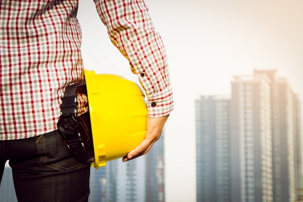 Trabalhador do engenheiro da mão que segura o capacete de segurança amarelo com a construção no fundo do local.