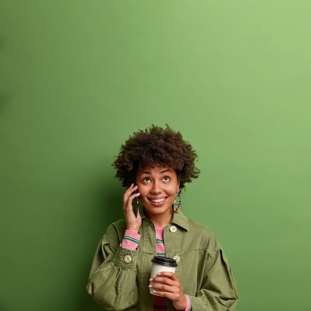 Trabalhadora étnica feliz trabalha produtivamente com um café energético, conversa ao telefone com um colega, olha para cima com um sorriso cheio de dentes, segura um copo descartável, espera alguém na reunião