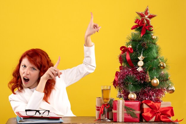 trabalhadora sentada atrás da mesa com os presentes de Natal e a árvore na mesa amarela