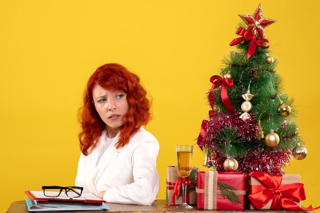 trabalhadora sentada atrás da mesa com presentes de Natal e árvore em amarelo