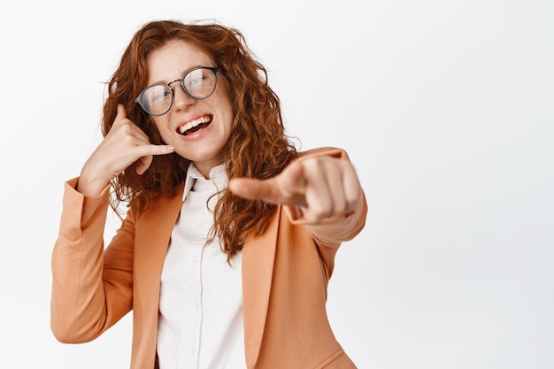 Trabalhadores de escritório garota ruiva sorridente de terno apontando o dedo para a câmera mostrando gesto de celular pedindo para ligar para ela recrutando você de pé sobre fundo branco