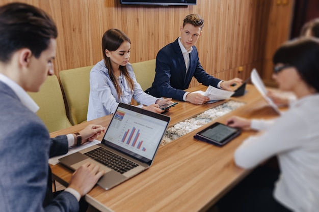 Trabalhadores de escritório realizar uma reunião em uma mesa para laptops, tablets e papéis, no fundo um grande aparelho de TV em uma parede de madeira