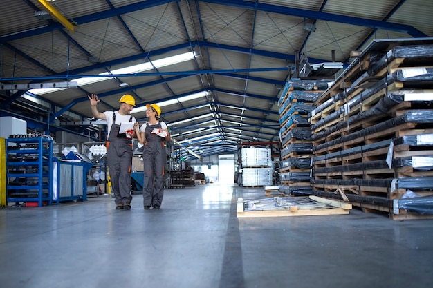 Trabalhadores de fábrica com roupas de trabalho e capacetes amarelos caminhando pelo hall de produção industrial e compartilhando ideias sobre organização