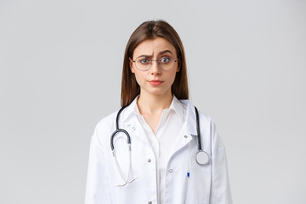 Trabalhadores de saúde, medicina, seguro e conceito de pandemia covid-19. Médica cética e confusa de uniforme branco, terno médico e óculos, sobrancelha levantada em julgamento, sorriso irônico