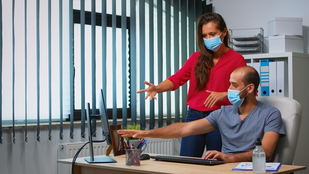 Trabalhadores falando usando máscaras de proteção na sala de escritório durante o coronavírus. Equipe no novo espaço de trabalho normal de escritório em empresa pessoal corporativa digitando no teclado do computador e apontando para a área de trabalho