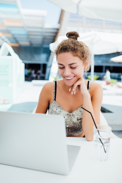 Trabalhar ao ar livre. Mulher jovem e bonita com chapéu descolado, trabalhando em um laptop e sorrindo enquanto está sentado ao ar livre