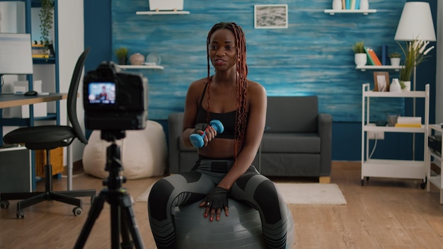 Foto grátis treinador atlético de mulher alongando os músculos do corpo, gravando o treino de ioga matinal com halteres na sala de estar. adulto slim fit vestindo roupas esportivas, filmando tutorial de ginástica, sentado na bola suíça