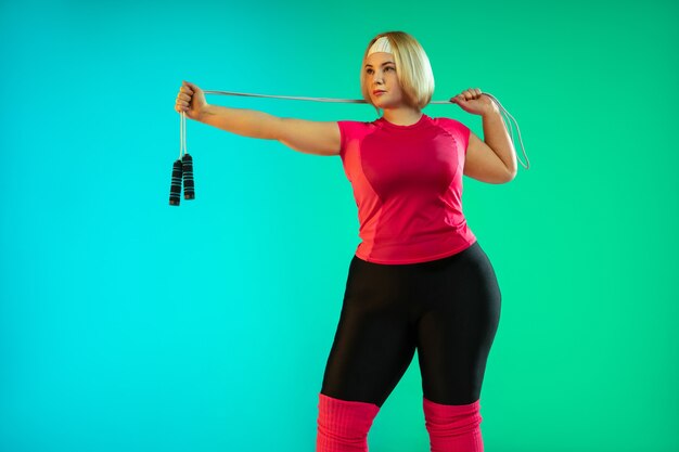 Treinamento do jovem modelo feminino caucasiano plus size em fundo gradiente verde em luz de néon. Fazendo exercícios de treino com corda de pular. Conceito de esporte, estilo de vida saudável, corpo positivo, igualdade.