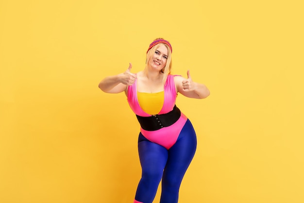 Foto grátis treinamento do modelo feminino jovem caucasiano plus size na parede amarela. copyspace. conceito de esporte, estilo de vida saudável, corpo positivo, moda, estilo. mulher elegante sorrindo com polegares para cima.