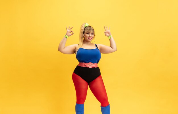 Treinamento do modelo feminino jovem caucasiano plus size na parede amarela. Mulher elegante com roupas brilhantes. Copyspace. Conceito de esporte, estilo de vida saudável, corpo positivo, moda. Sorrindo, mostrando-se bem.